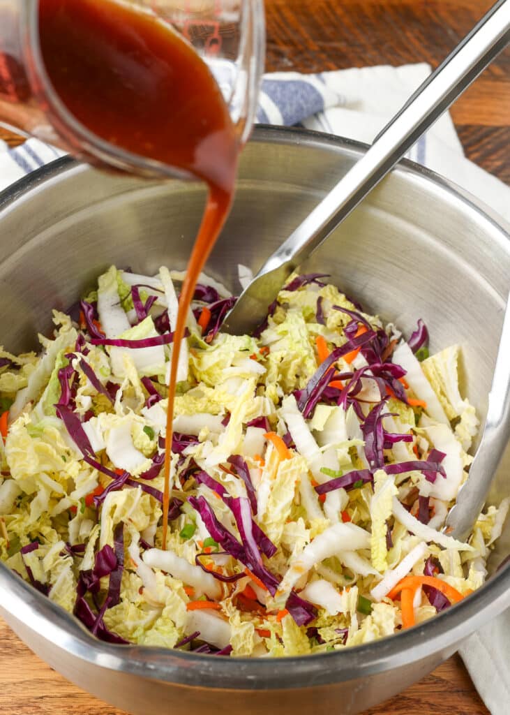 multi color Asian cabbage salad in a mixing bowl with tongs on a wooden surface with a blue and white linen