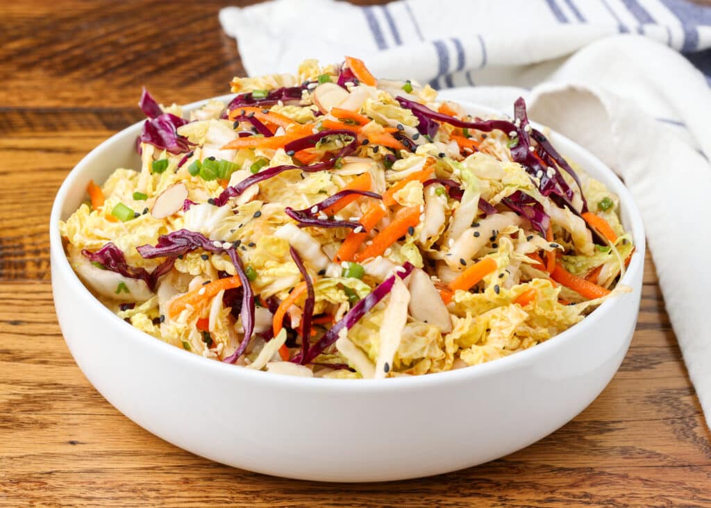 multi color Asian cabbage salad in a white bowl on a wooden surface with a blue and white linen