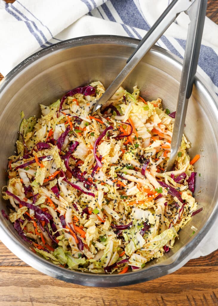 multi color Asian cabbage salad in a mixing bowl with tongs on a wooden surface with a blue and white linen
