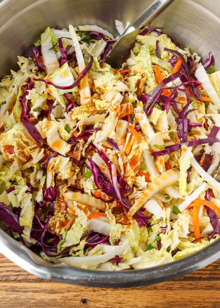 multi color Asian cabbage salad in a mixing bowl with tongs on a wooden surface with a blue and white linen