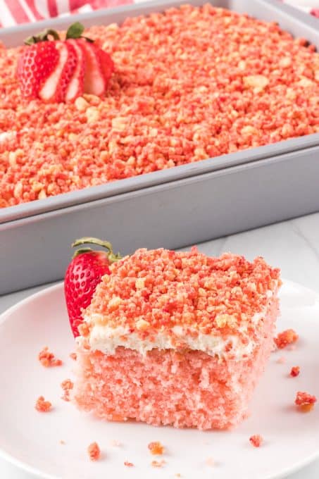 A pink strawberry flavored cake on a plate with a strawberry in front of a cake pan.