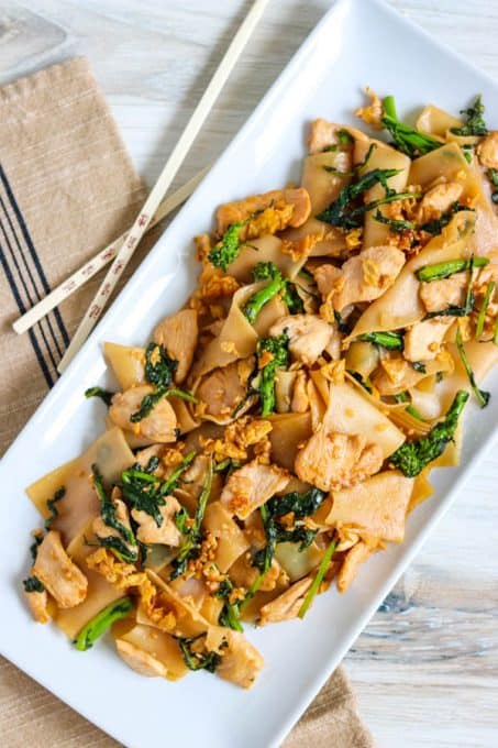Chicken, wide rice noodles and broccoli rabe on a plate with chopsticks.