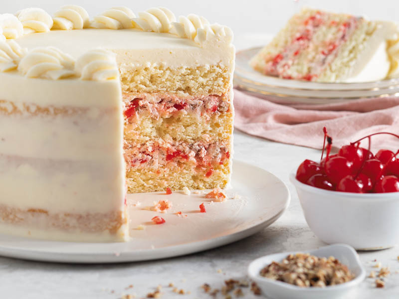 cherry-pecan cake on a serving plate with a slice of cake on another plate and a bowl of cherries and a bowl of pecans