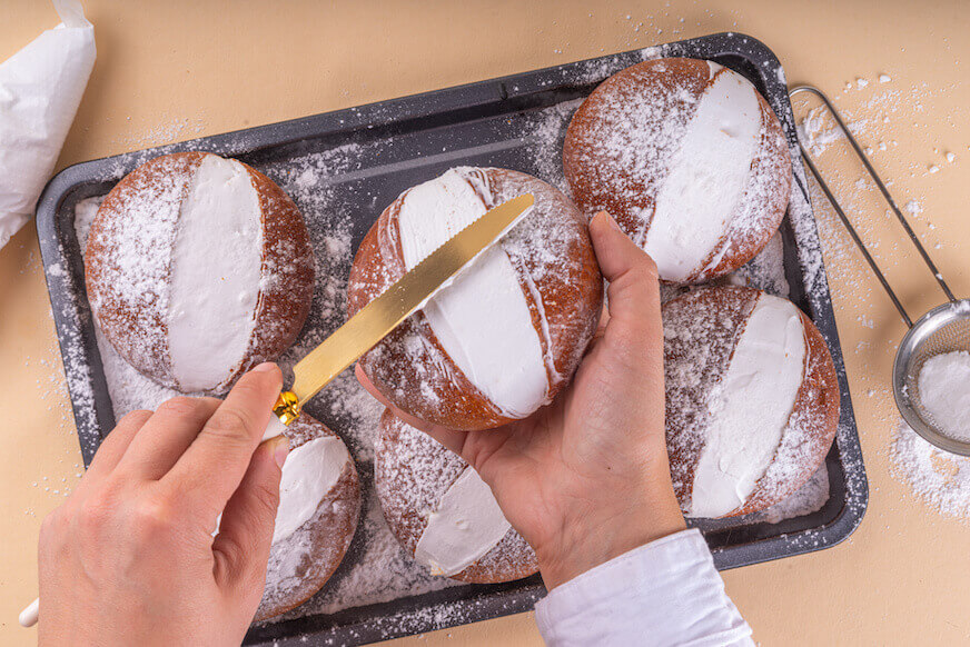 An overhead view of a pan of maritozzi, with a pair of hands holding one and smoothing out the whipped cream filling with a butter knife.