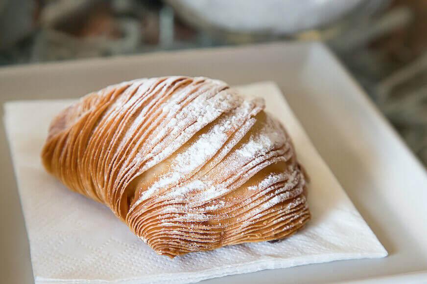 A sfogliatella sits on a white plate and is dusted with confectioner's sugar.