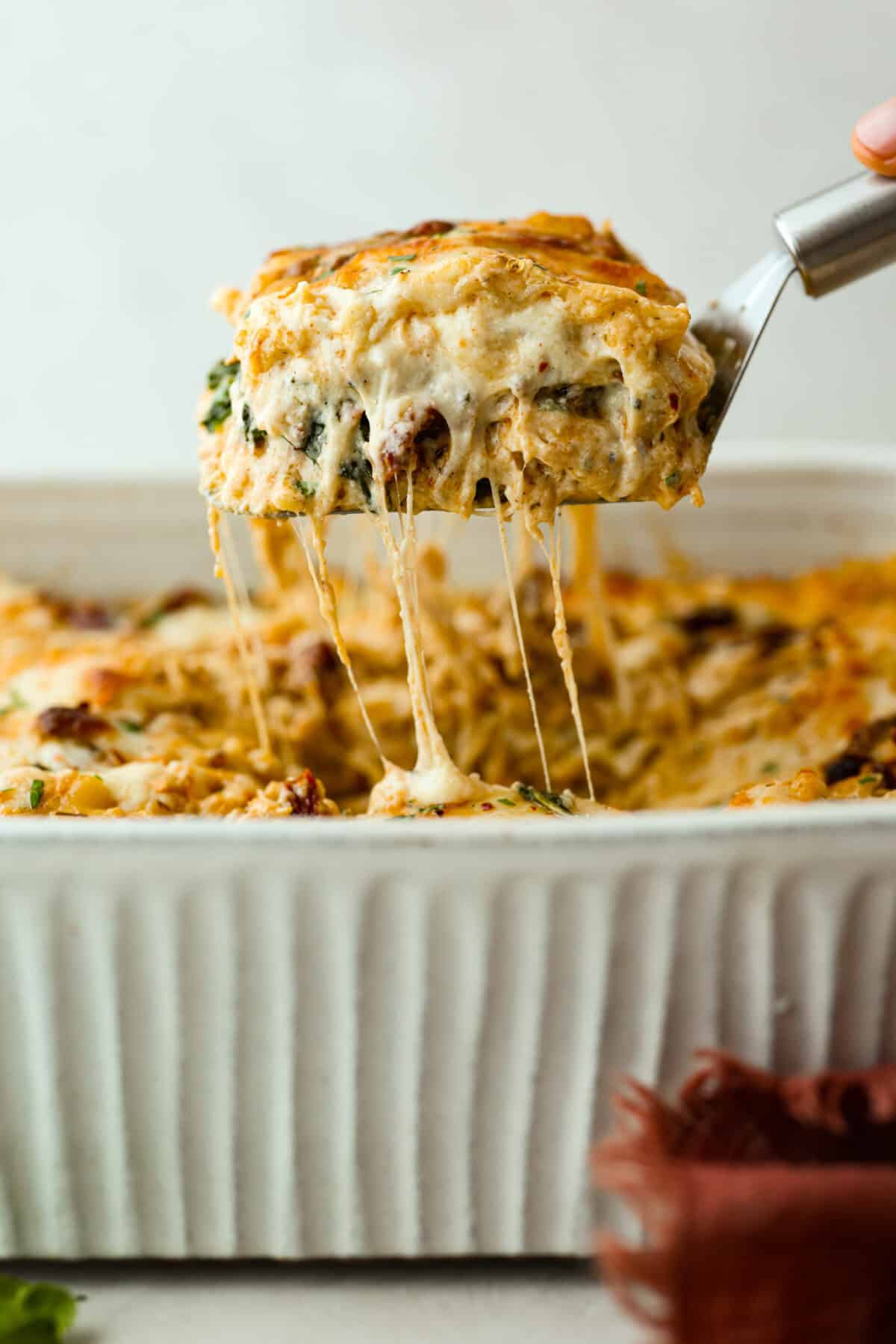Side shot of someone pulling a slice of marry me lasagna out of the pan with cheesy sauciness stretching from the main casserole. 