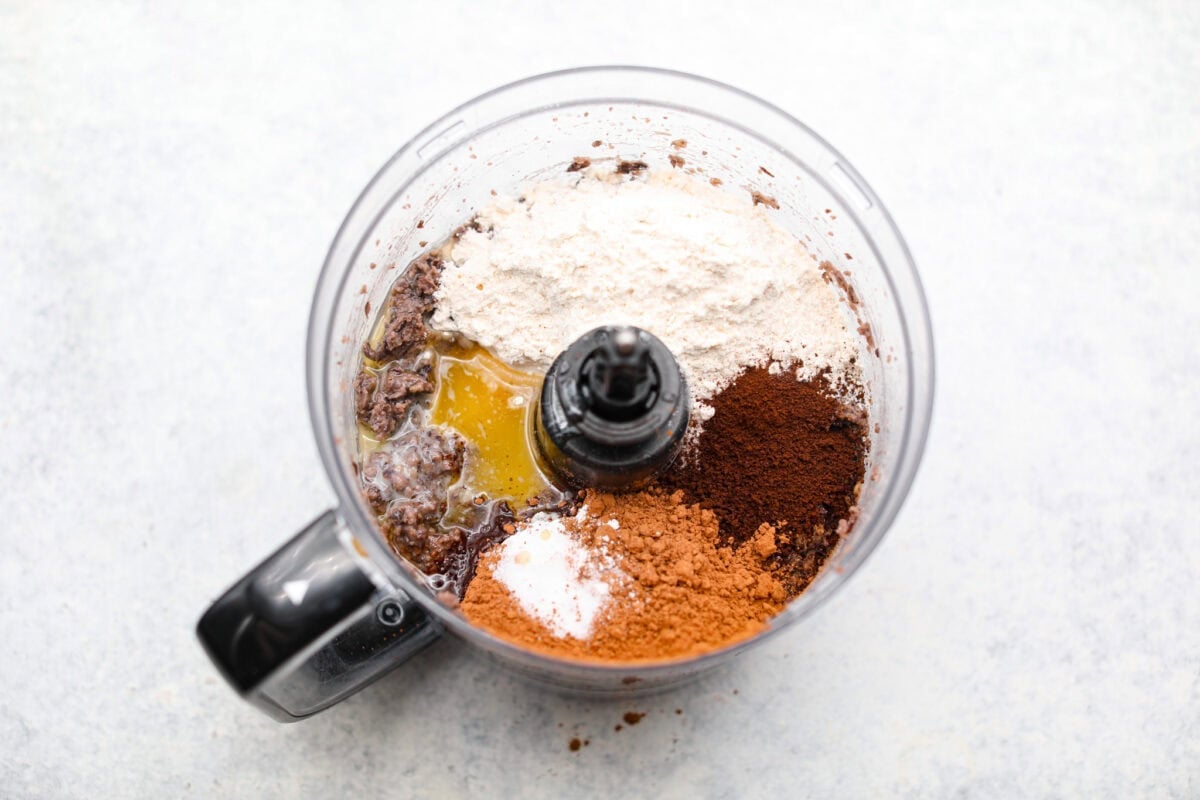 Overhead shot of the coco powder, instant coffee, egg sugar, melted butter, salt, vanilla extract, baking powder and flour added to the black beans in the food processor. 