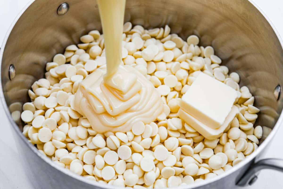 Someone pouring sweetened condensed milk into a sauce pan with white chocolate and butter. 