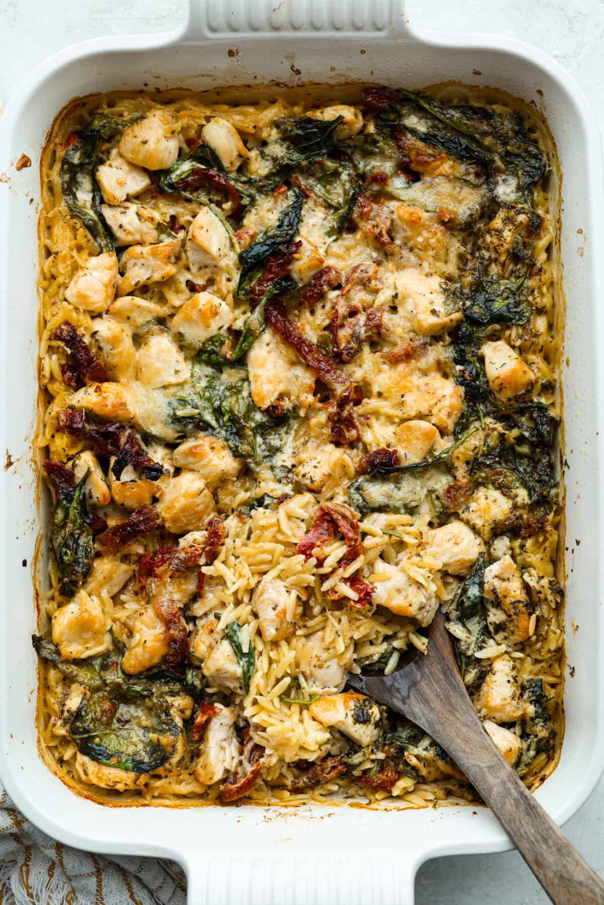 Tuscan chicken orzo bake all cooked in a casserole dish with a wooden serving spoon.