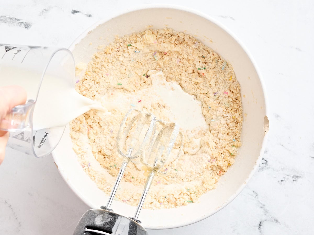 Milk being poured into a flour, butter, and egg mixture in a mixing bowl.