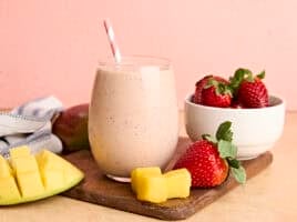 A glass of homemade strawberry mango smoothie with a straw, on a wooden cutting board with strawberries and mango chunks.