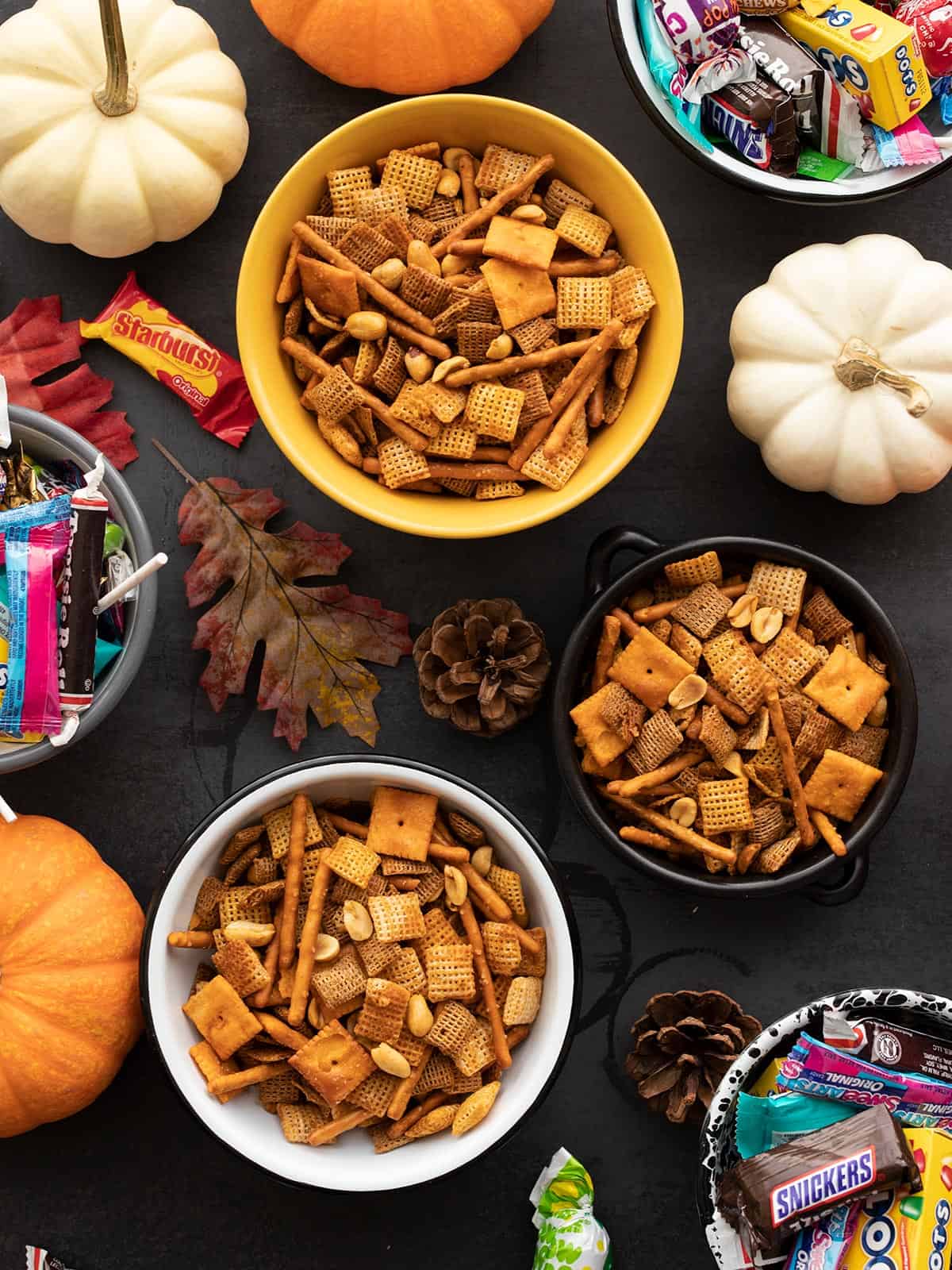 Three bowls of chex mix next to bowls of candy and pumpkins