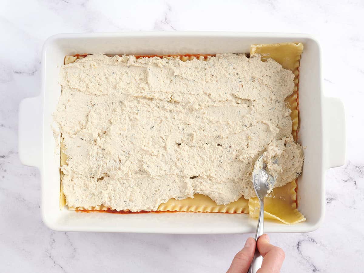 A hand using a spoon to spread tofu ricotta in a vegan lasagna.