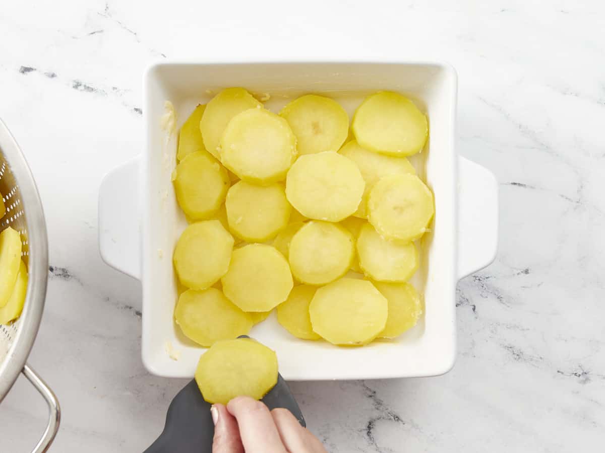 Overhead shot of scalloped potatoes being layered into a white casserole dish.