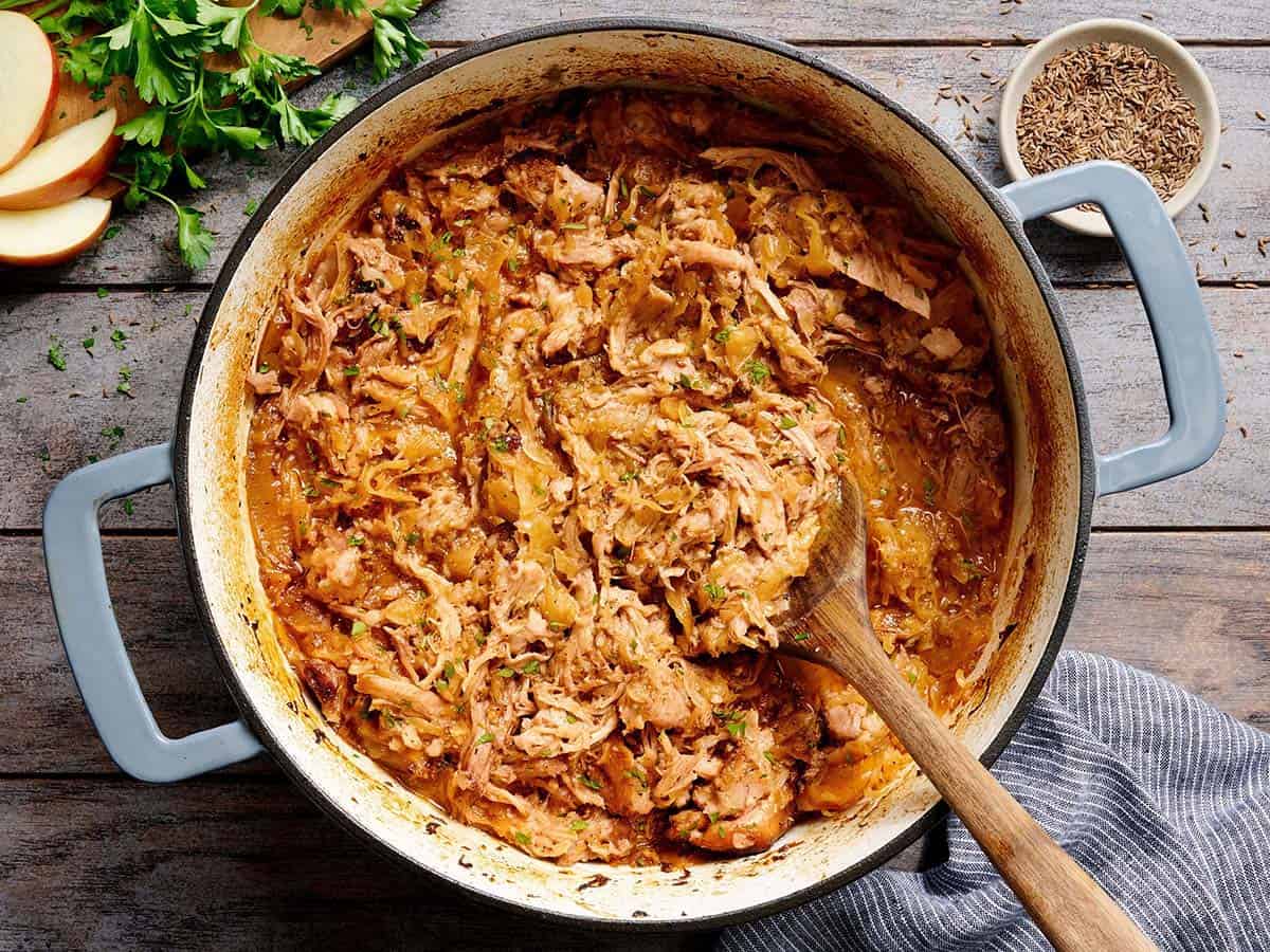 Overhead view of shredded pork and sauerkraut in a pot with a wooden spoon.