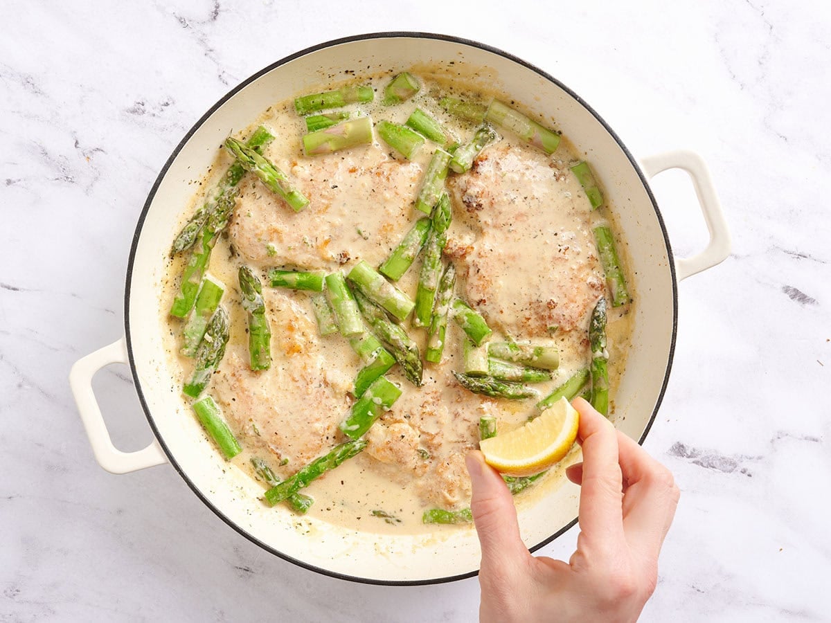 Hand squeezing a lemon over creamy asparagus chicken in a skillet.