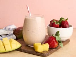 A glass of homemade strawberry mango smoothie with a straw, on a wooden cutting board with strawberries and mango chunks.