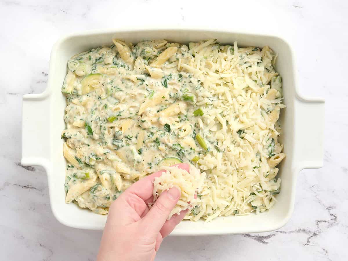 Pasta with spring veggies in a casserole dish being topped with mozzarella.