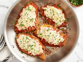 overhead view of 4 fried chicken breasts topped with marinara sauce, melted cheese, and parsley in a stainless skillet.