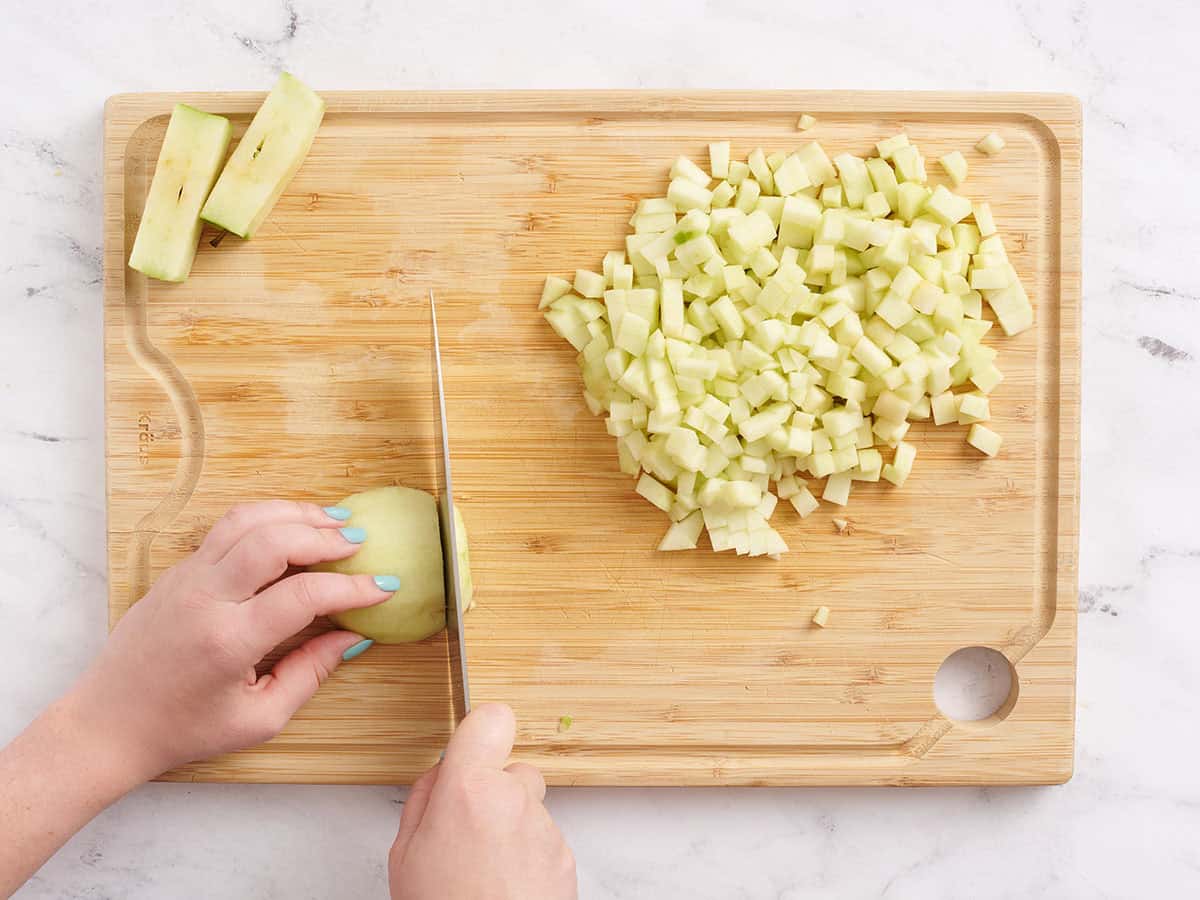A hand using a knife to dice an apple into small pieces on a wooden cutting board.