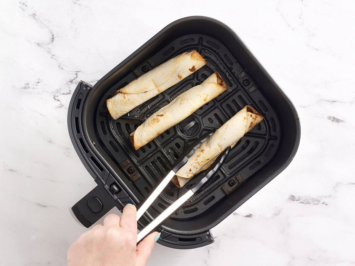 Kitchen tongs flipping over filled apple flautas in an air fryer basket.