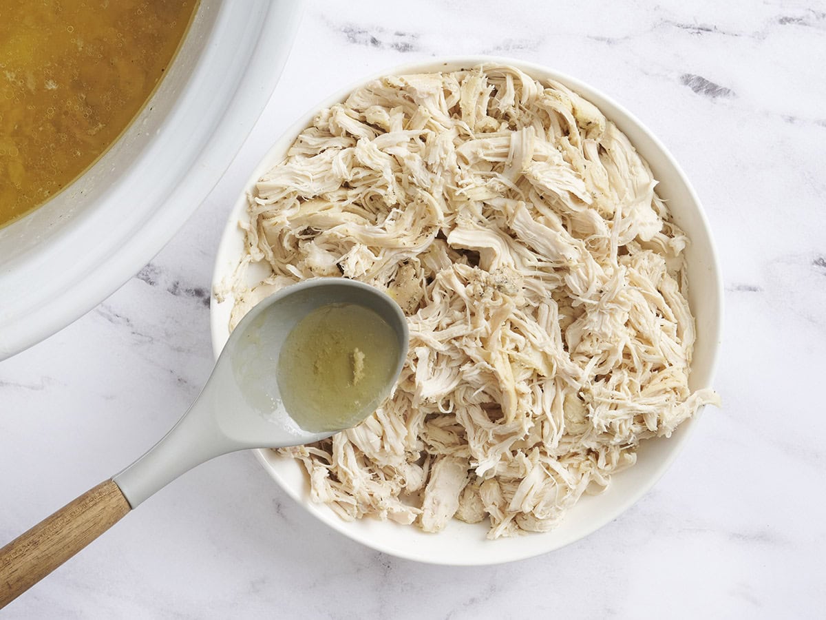 A spoonful of chicken broth being poured over shredded chicken on a plate.