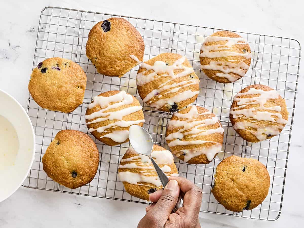 A hand drizzling a simple lemon glaze over lemon blueberry muffins.