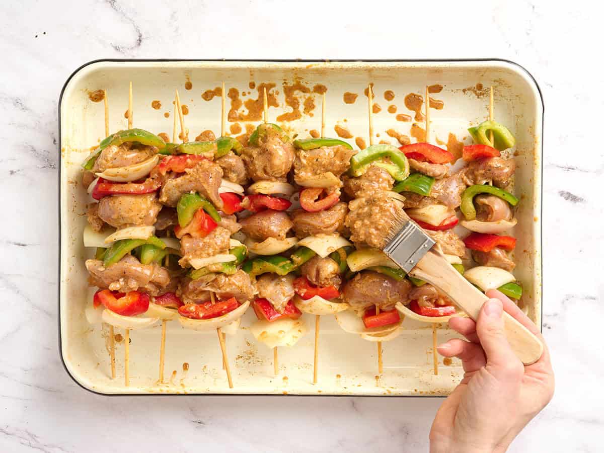 A hand brushing a marinade over chicken kebabs on a baking sheet.