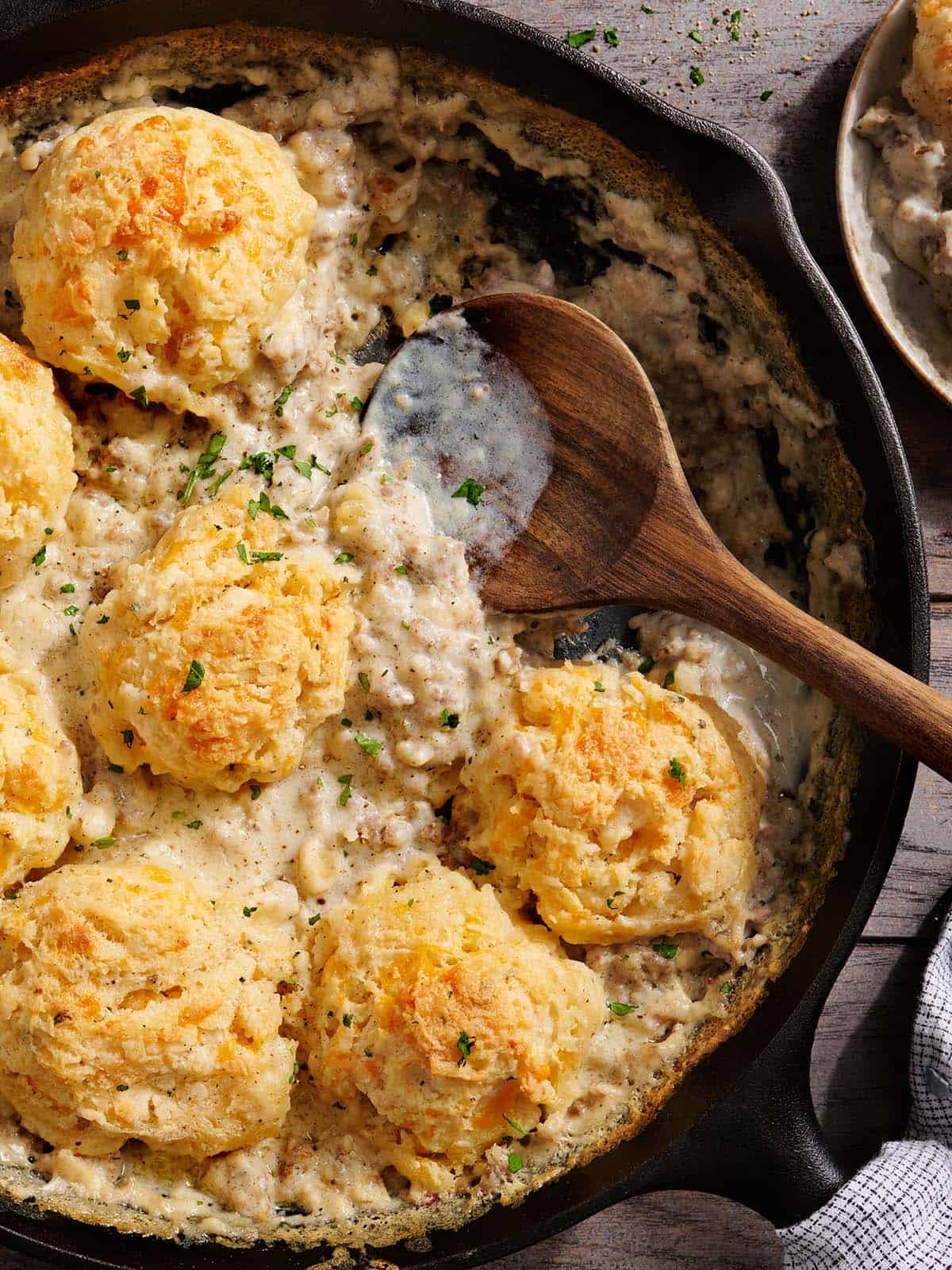 Overhead view of a biscuits and gravy bake in a skillet with a wooden spoon.
