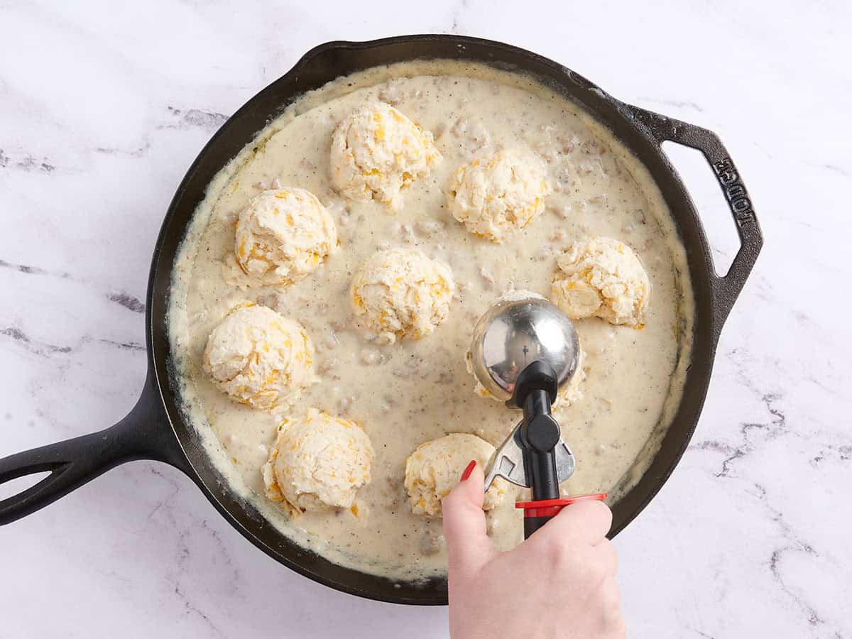 A hand using a scoop to portion biscuit dough over sausage gravy.