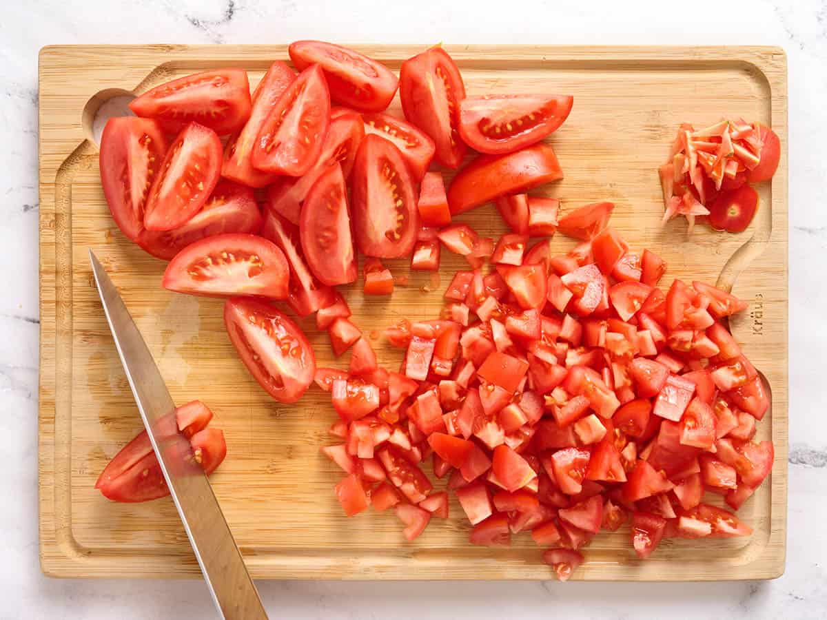 A knife chopping roma tomatoes on a wooden cutting board.
