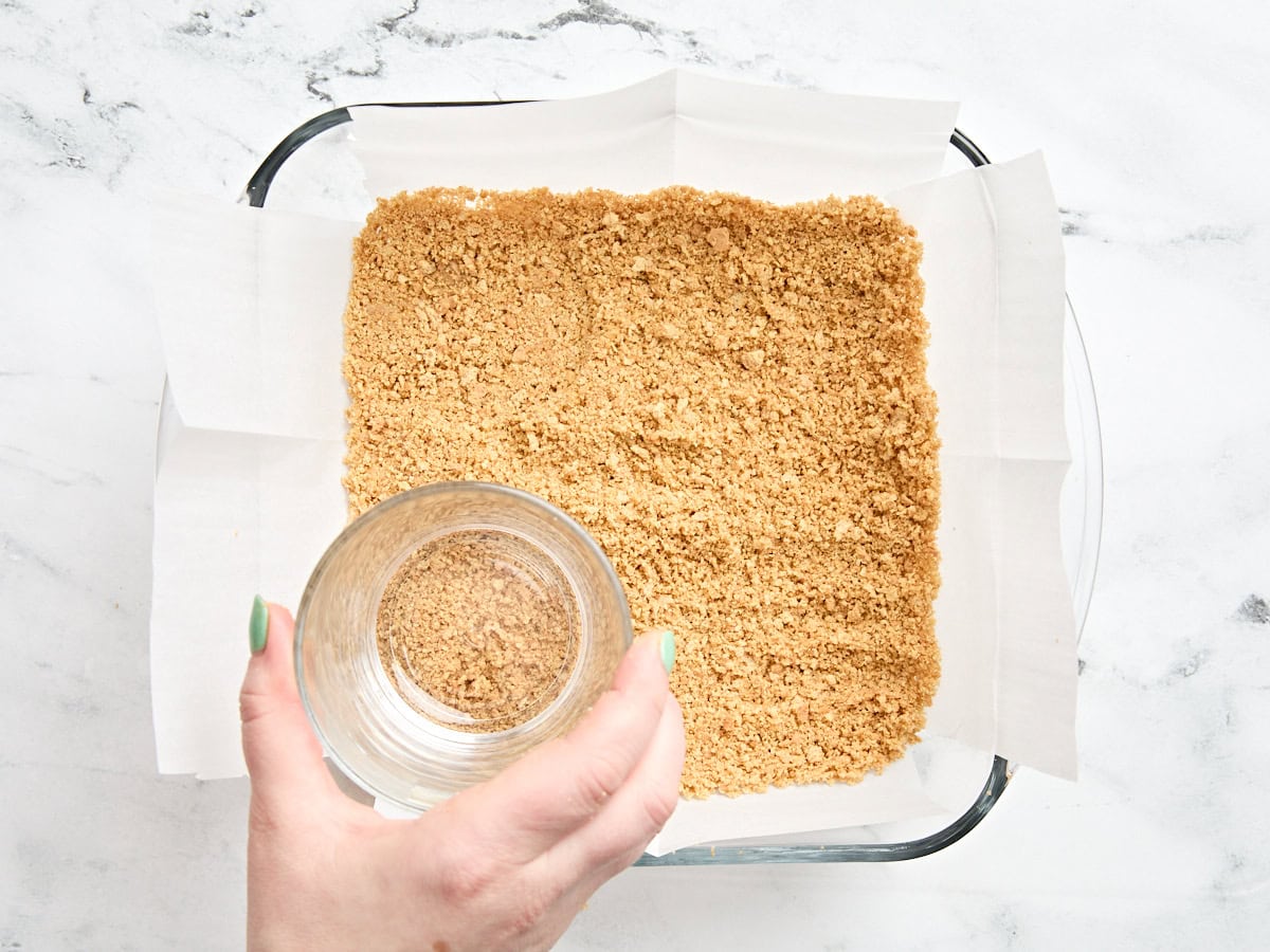 A hand using a glass to press graham cracker crumbs into a baking dish.