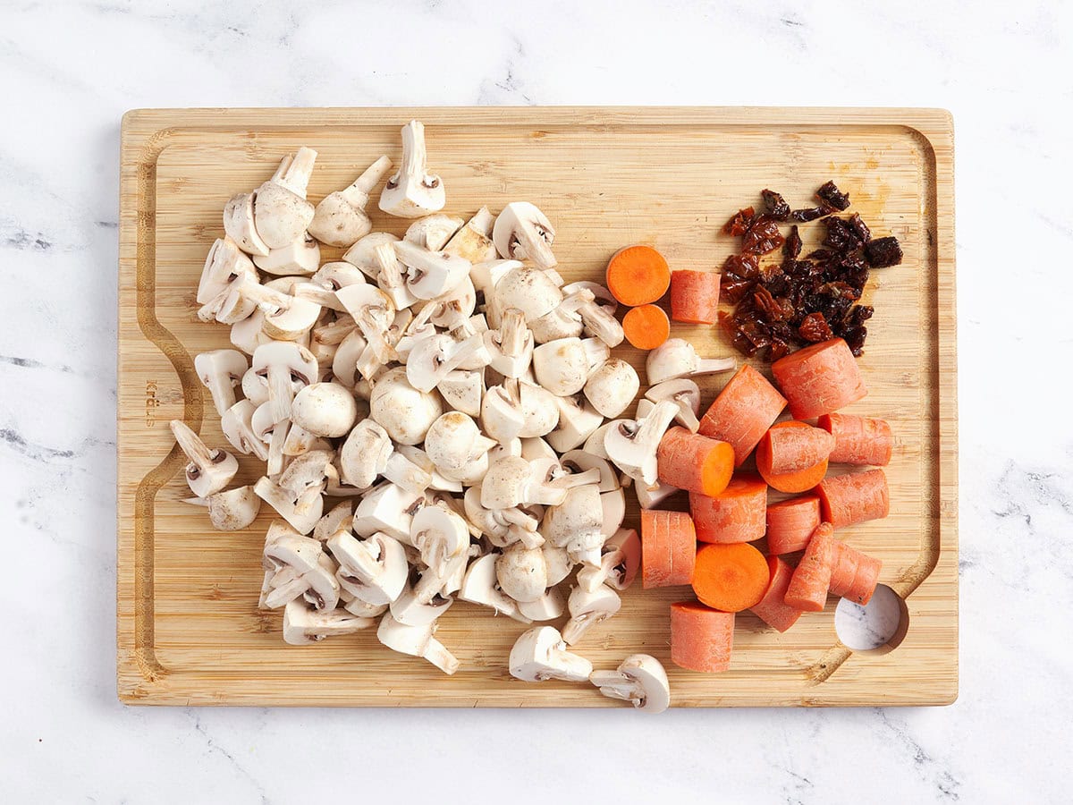 Chopped white mushrooms, carrots, and sun dried tomatoes on a wooden cutting board.