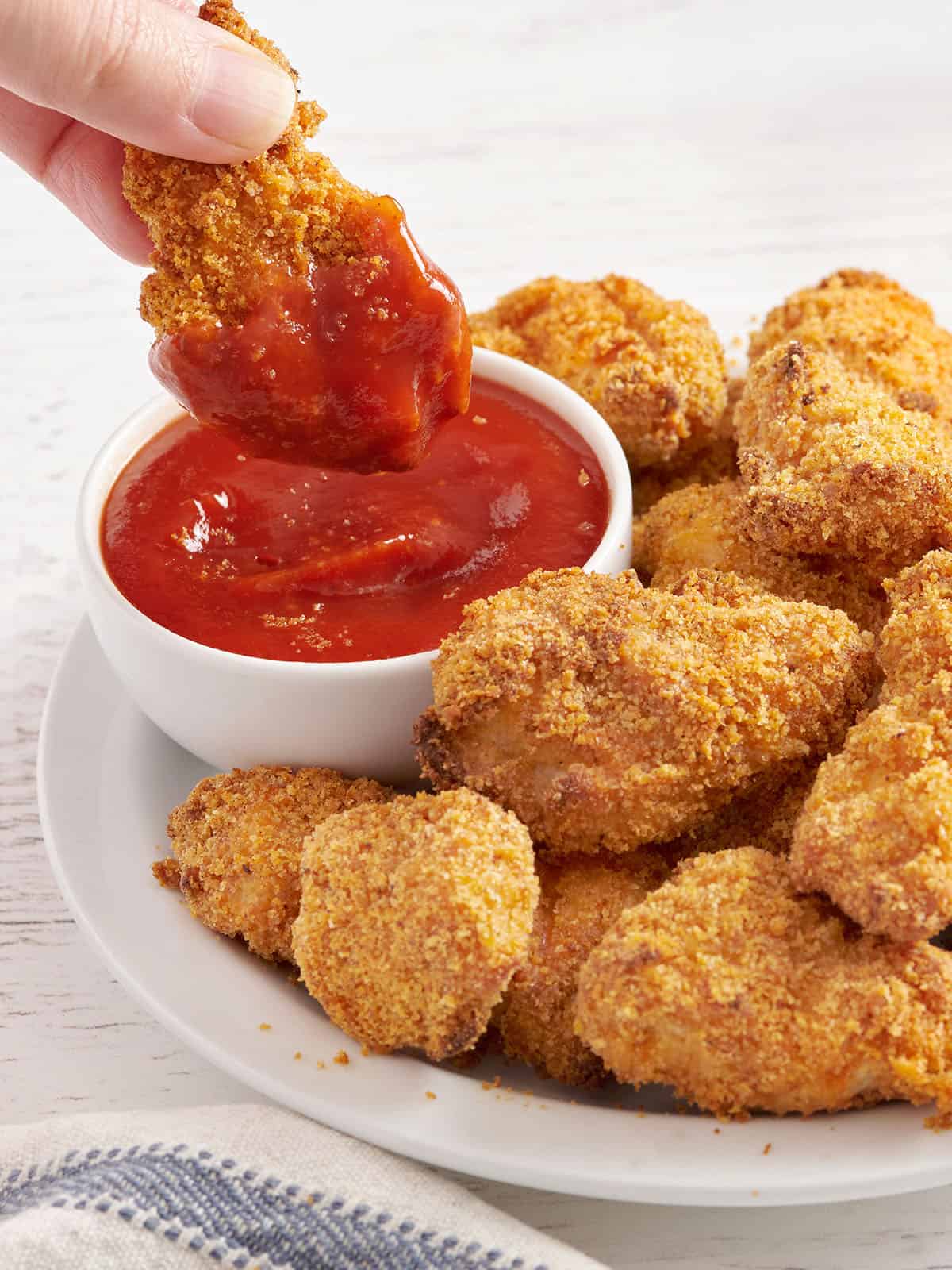 Side view of a plate of homemade chicken nuggets with a hand dipping a nugget into a pot of ketchup.