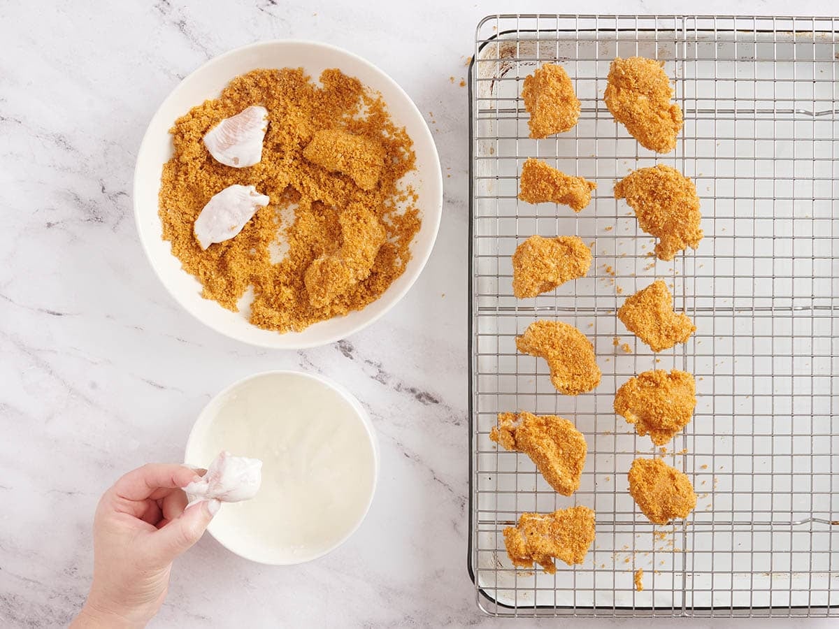 A hand dipping chicken in a yogurt mix, then in a breadcrumb mix and then lay on a wire rack.