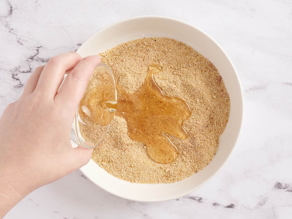 A hand pouring oil into a breadcrumb mixture for chicken nuggets.