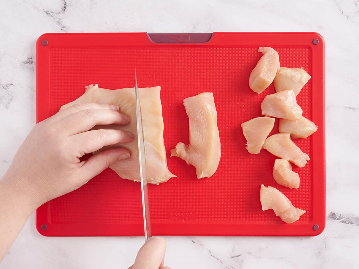 A hand using a knife to cut chicken breast into chunks.