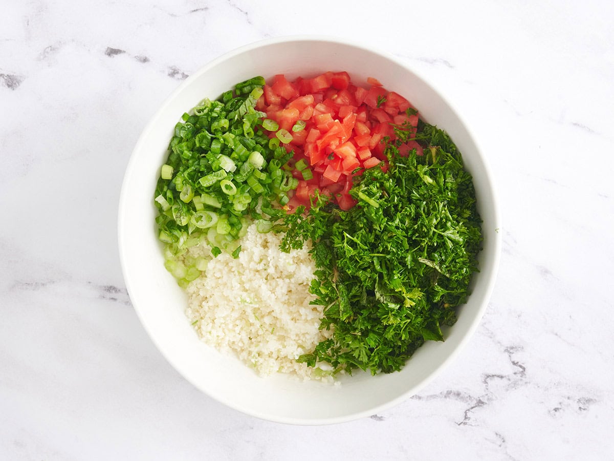 Diced green onions, riced cauliflower, tomatoes and herbs in a bowl.