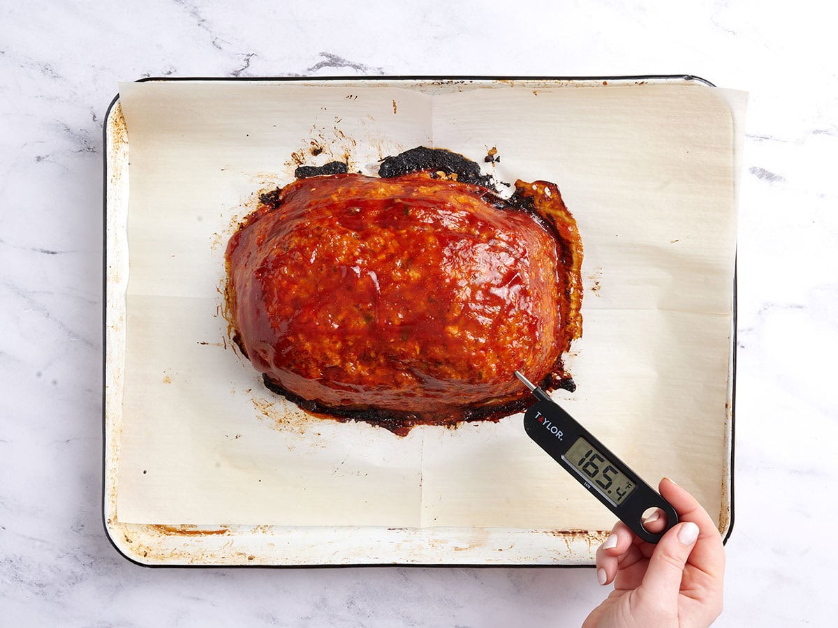 A thermometer reading 165&deg;F being stuck into the chicken meatloaf on a white baking sheet lined with parchment paper. 