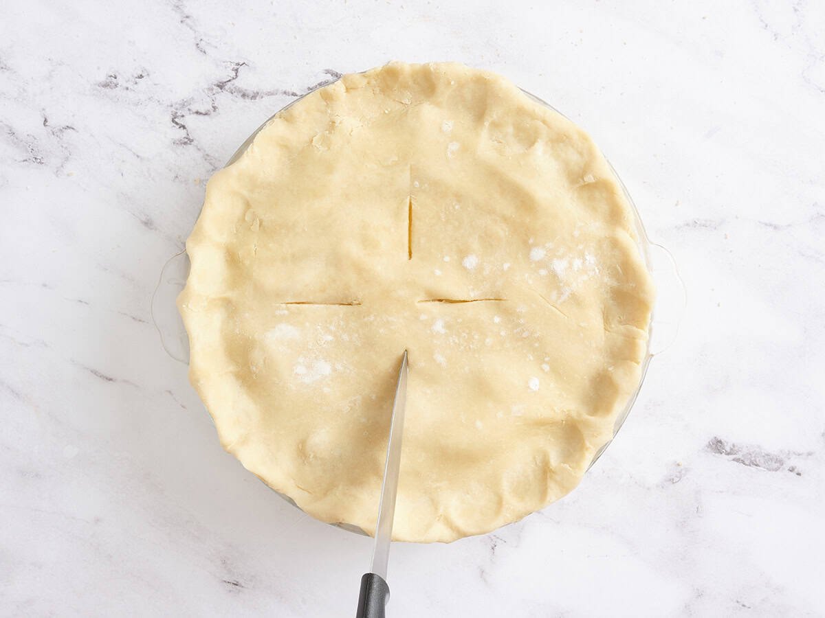 A knife slicing slits into the top of a homemade apple pie.