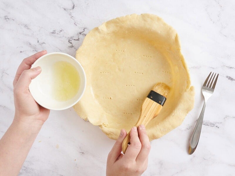 Egg wash being brushed onto a pie crust in a pie dish.