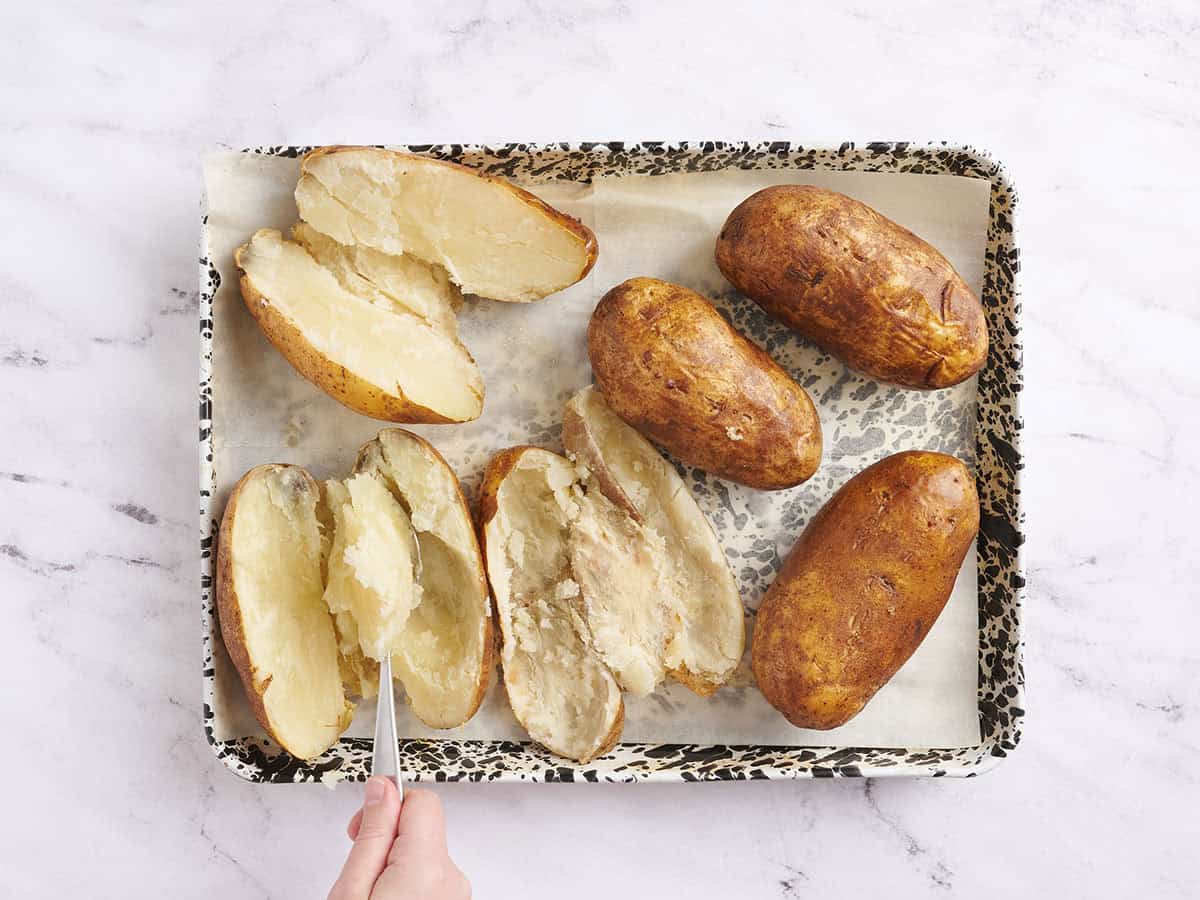 A hand using a spoon to scoop out the insides of baked potatoes on a baking sheet.