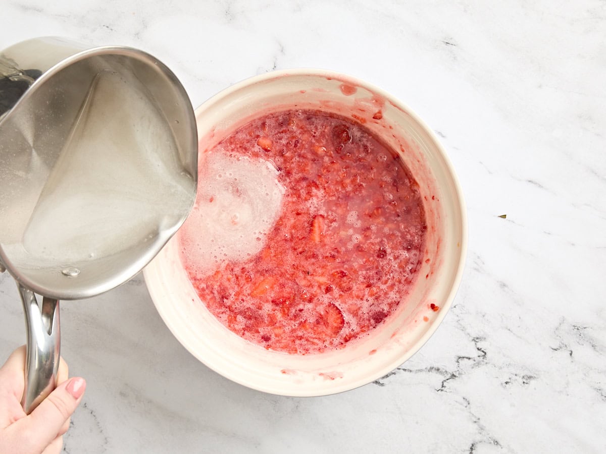 Boiled fruit pectin mixture being poured into a bowl of chopped strawberries and sugar.