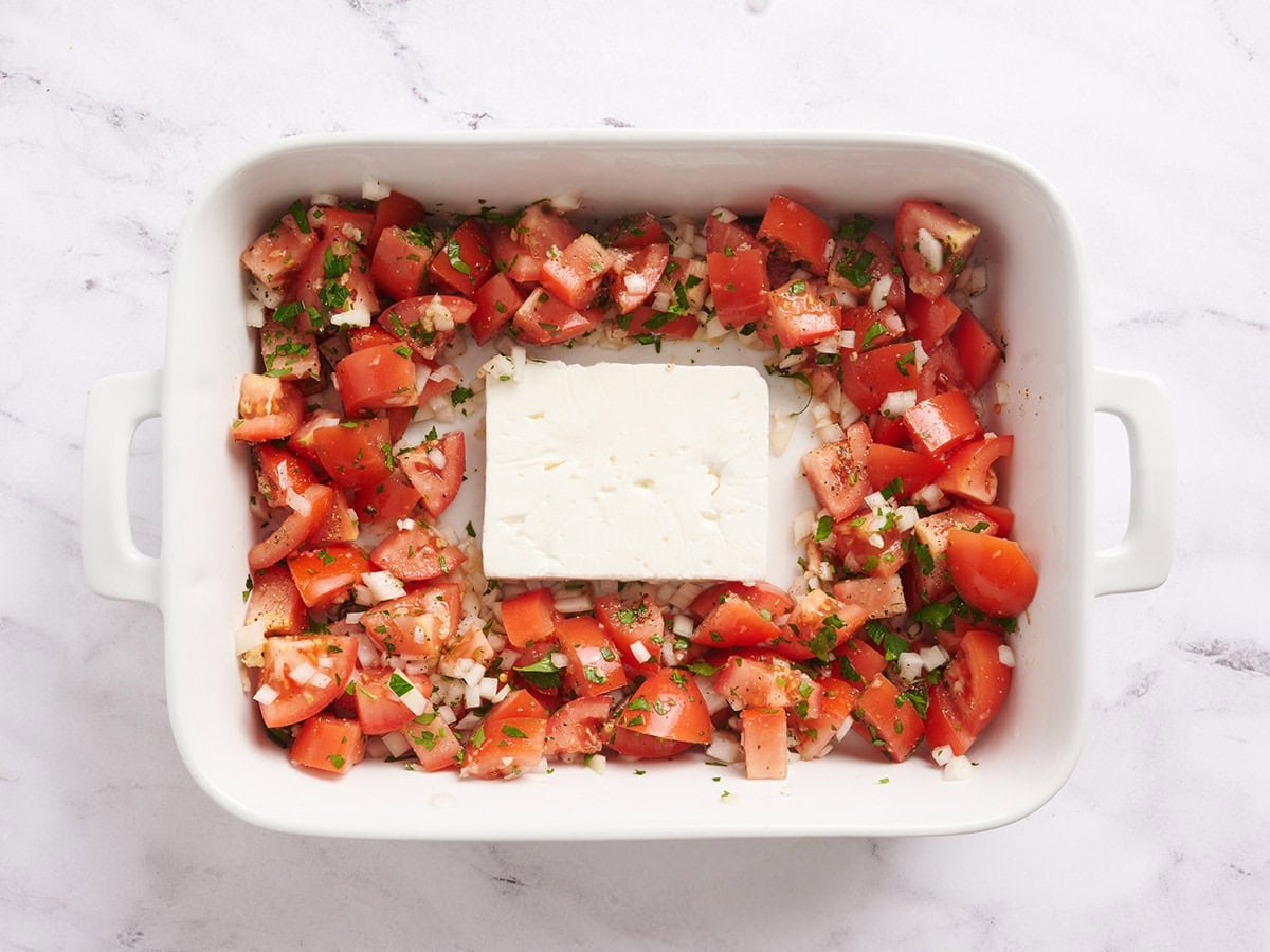 Diced tomatoes and onions in a baking dish with a block of feta in the middle.