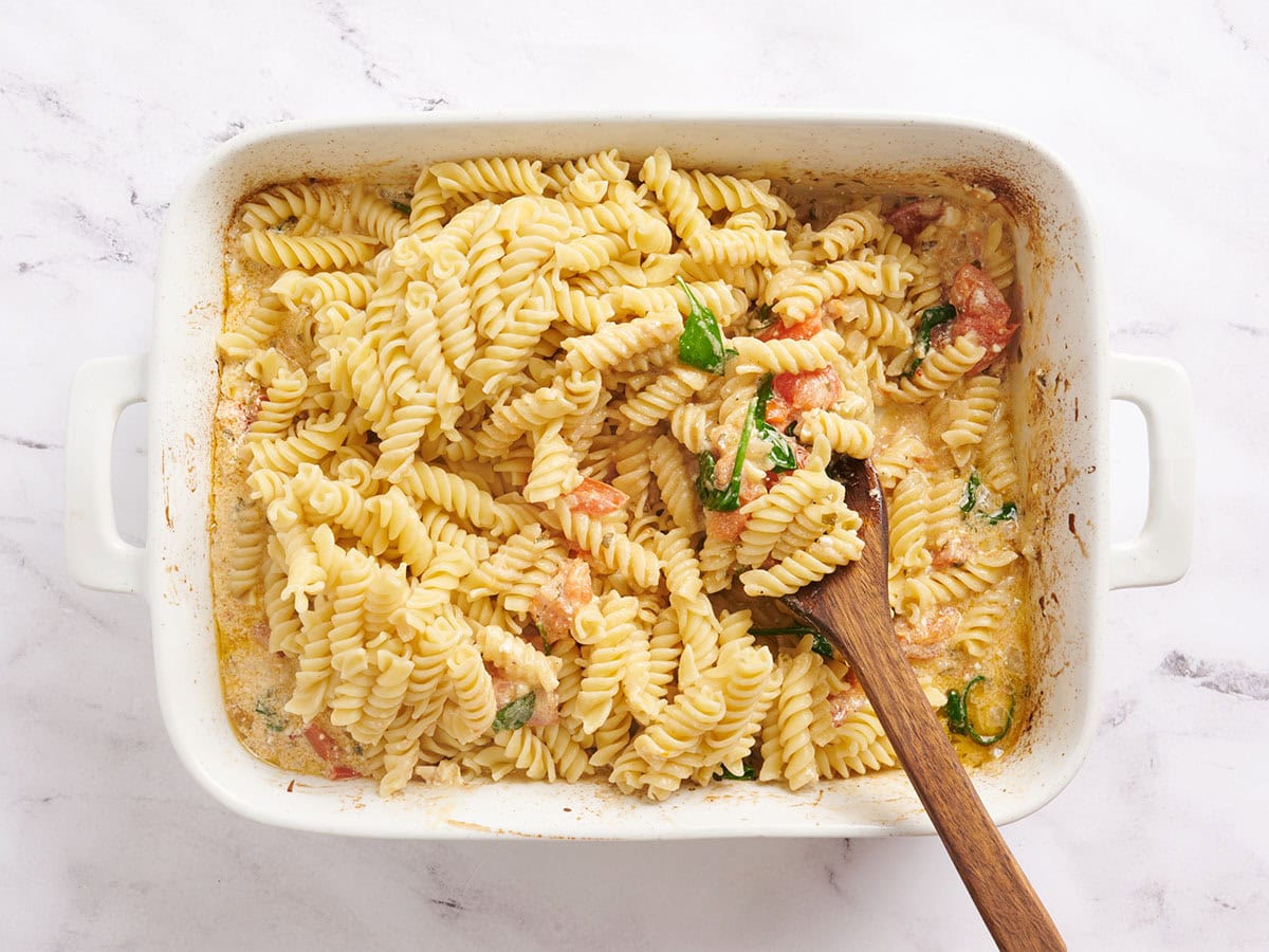 Rotini pasta being mixed into roasted tomatoes, onions, and feta cheese.