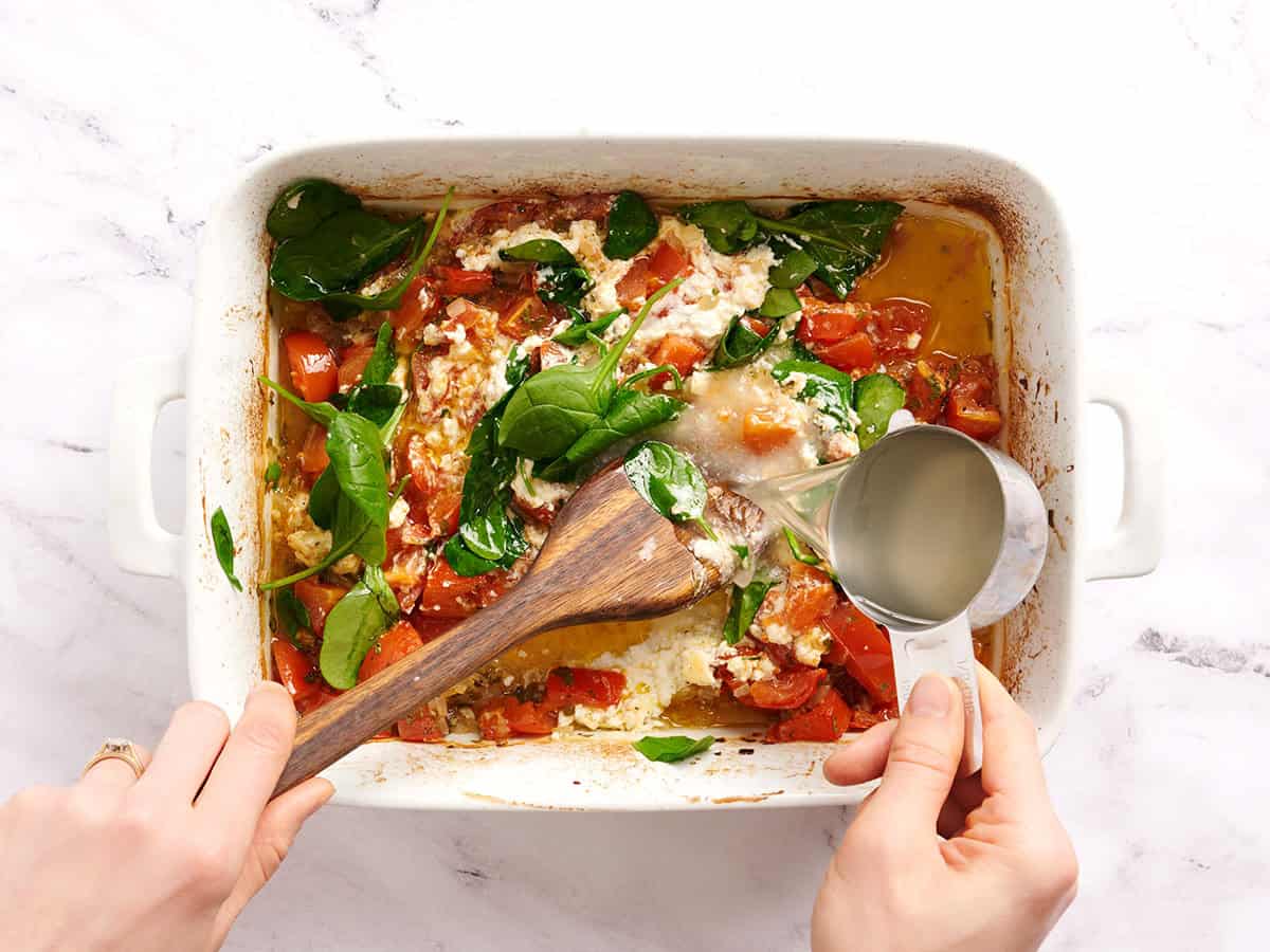 A hand pouring in reserved pasta water into a baking dish of baked tomatoes and feta cheese.