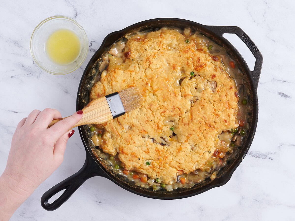 A hand brushing melted butter on a chicken cobbler.