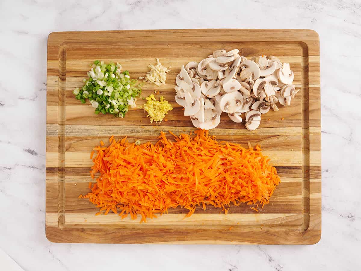 Sliced mushrooms, grated carrots, minced green onions, garlic and ginger on a wooden cutting board.