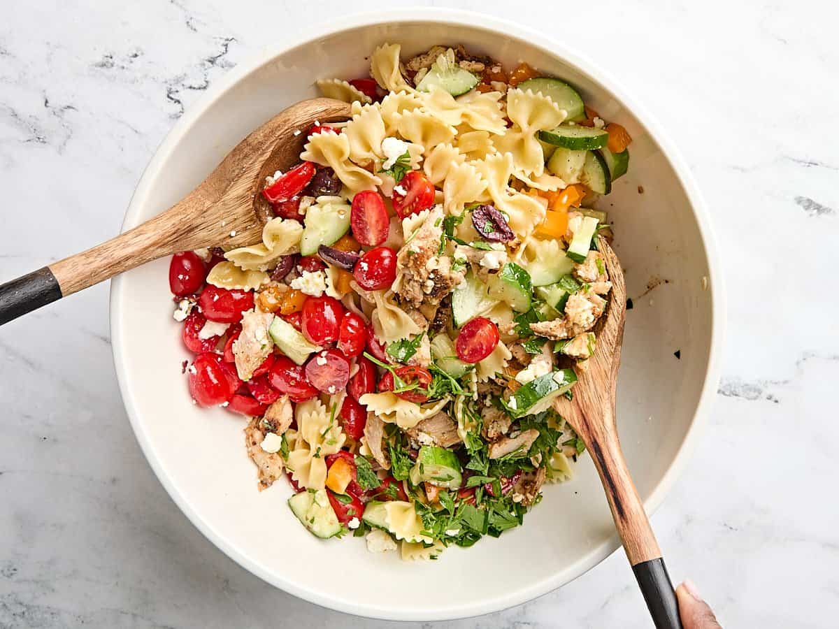 Two wooden spoons tossing a greek chicken salad in a bowl.