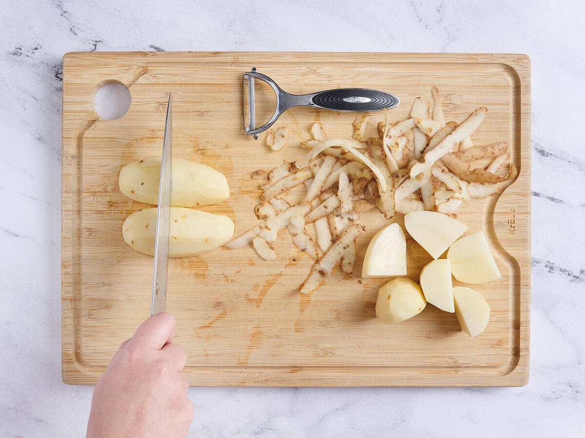 Peeled potatoes on a wooden cutting board being sliced with a knife.